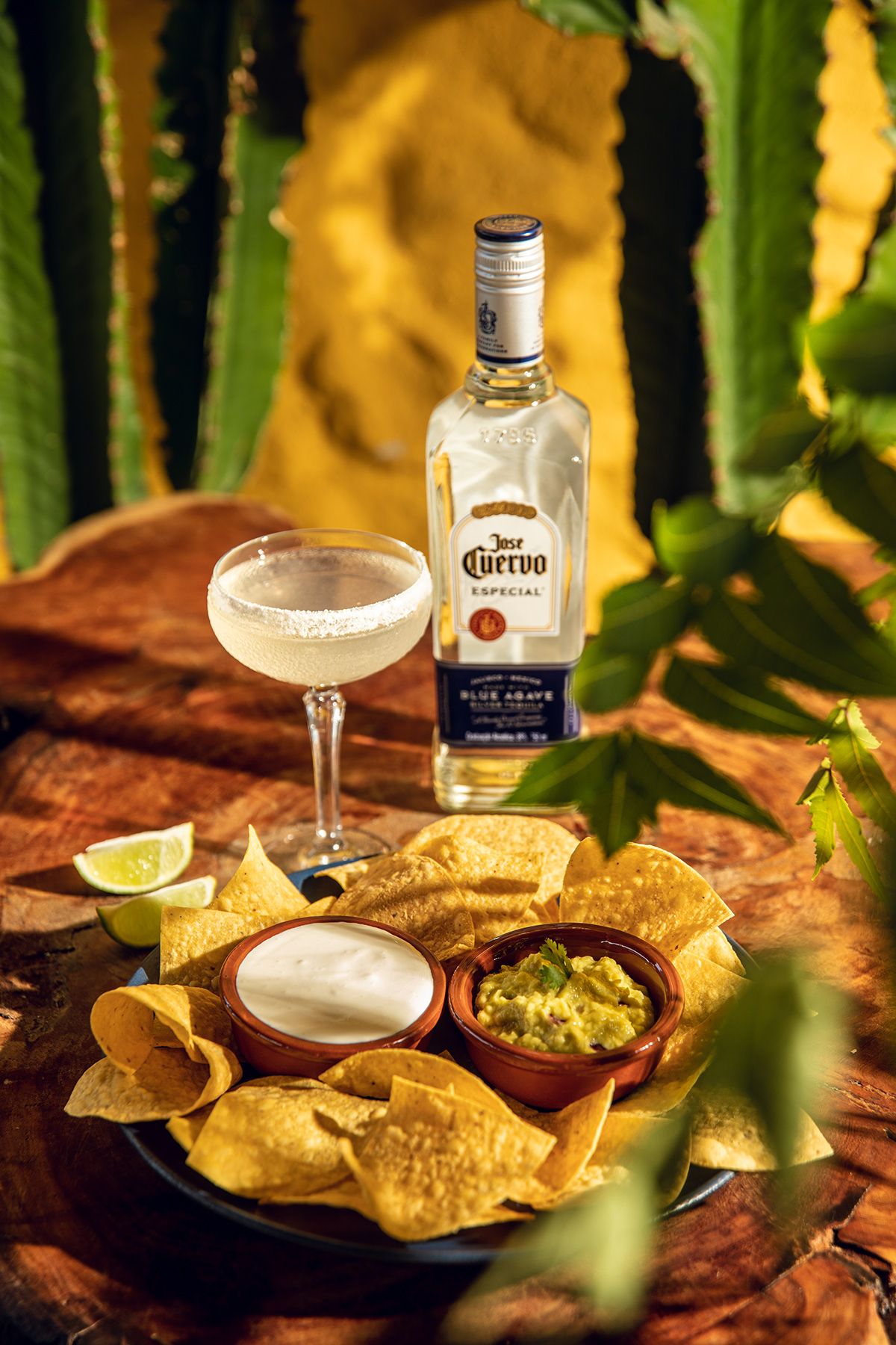 Plate of tortilla chips with bowls of guacamole and sour cream, lime wedges, a margarita glass with salt rim, and a bottle of Jose Cuervo Especial tequila on a wooden table.