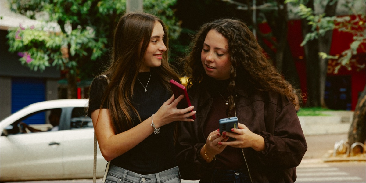 Duas amigas sorrindo enquanto olham seus celulares na rua