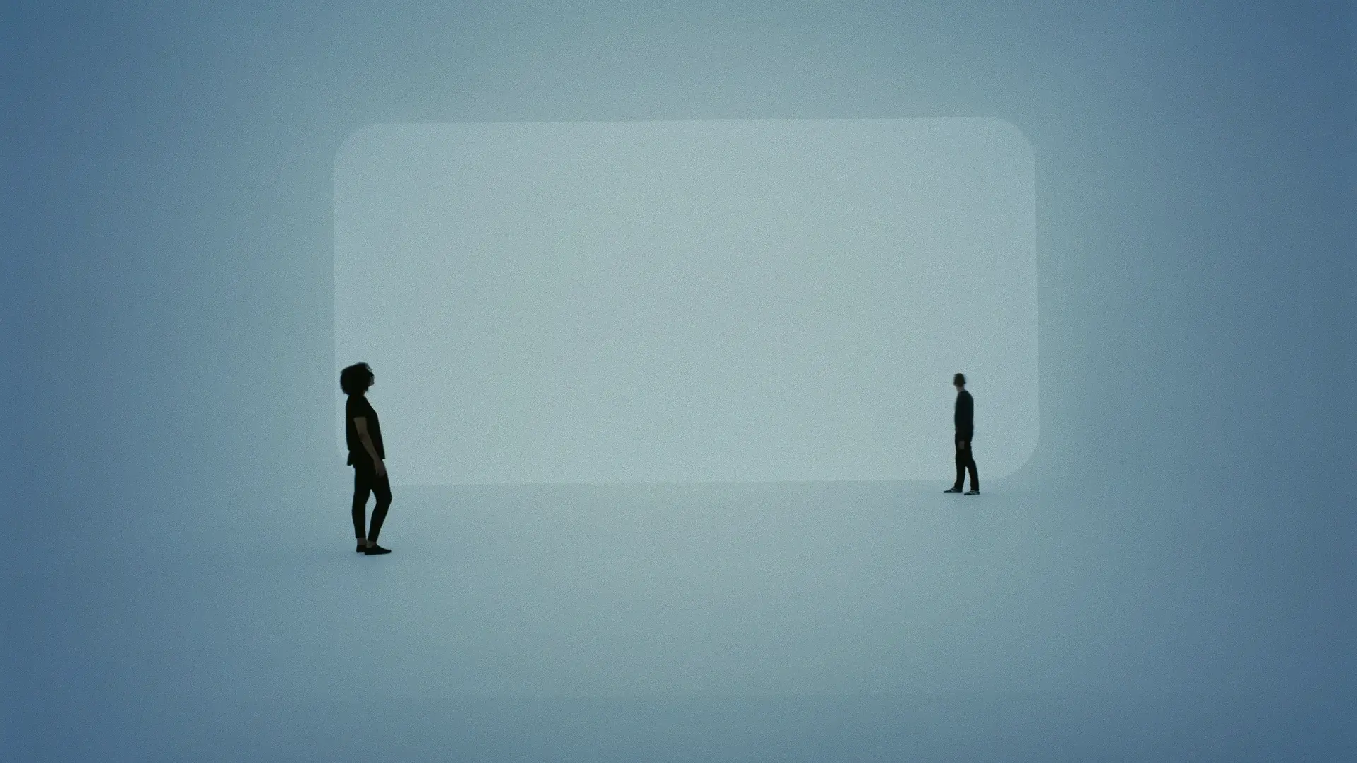 Two people standing facing each other in a large, empty, softly lit room with a rounded rectangle light shape on the wall behind them.
