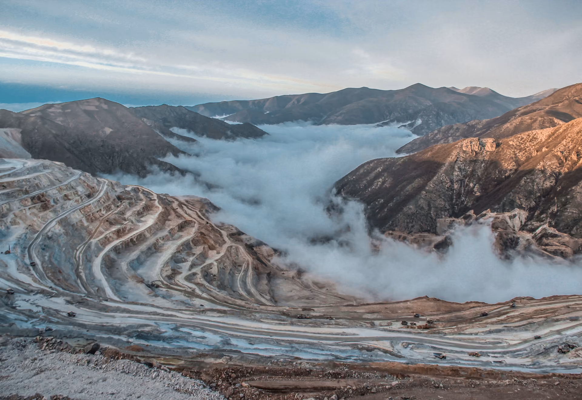 Overhead view of a terraced open-pit mine surrounded by mountains and a low cloud layer.