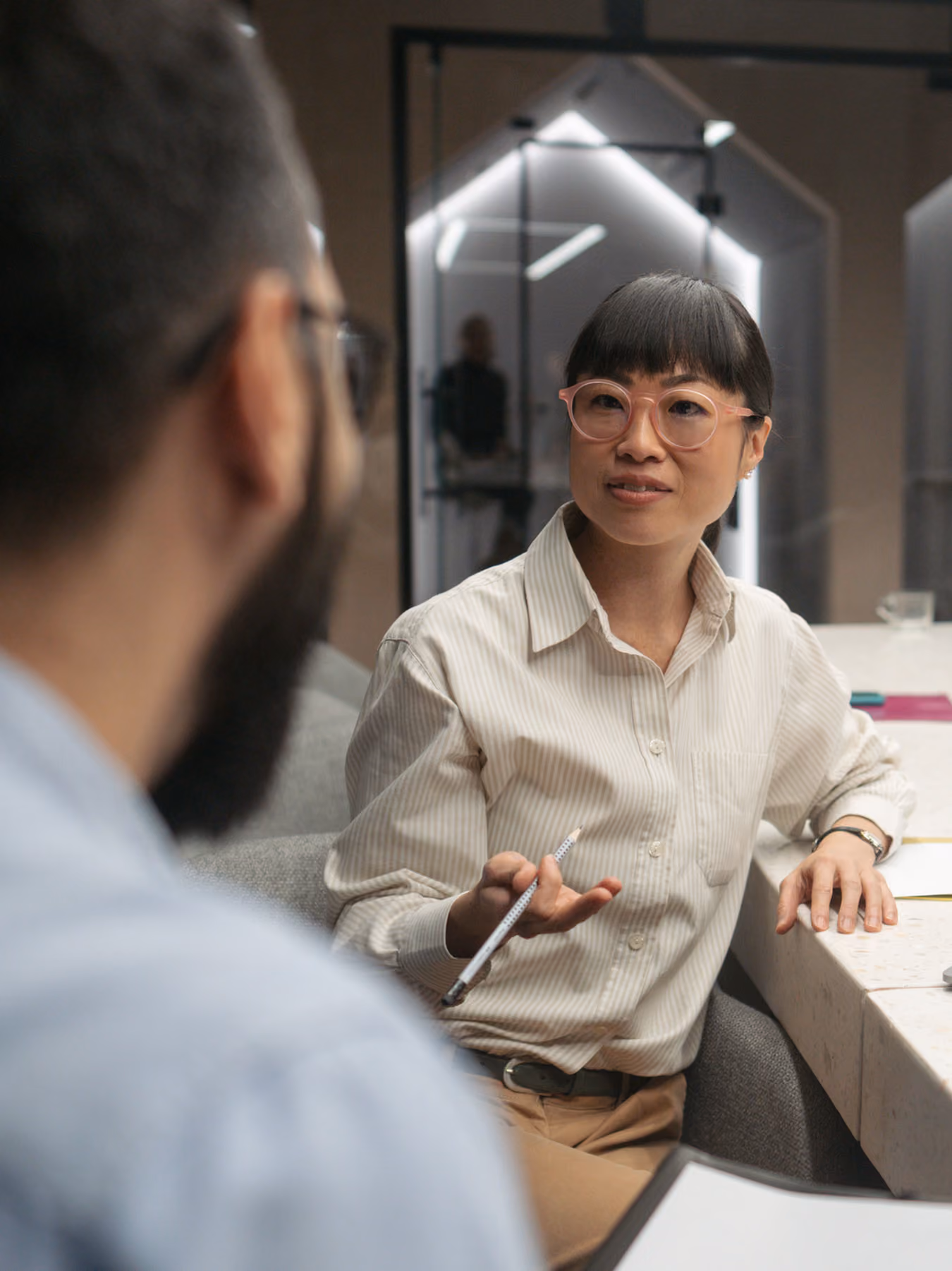 Woman with pink glasses and striped blouse talking and gesturing with a pencil at a table with a man.