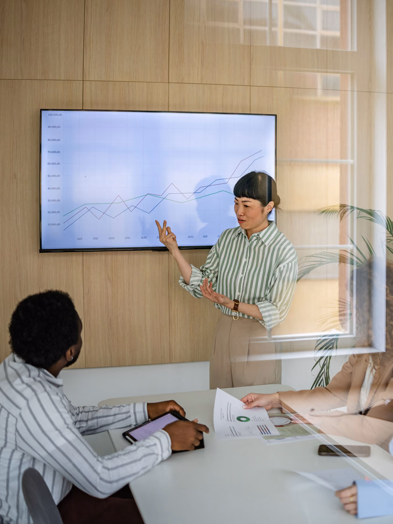 Woman presenting line graph on screen to colleagues seated at table with documents and tablet.