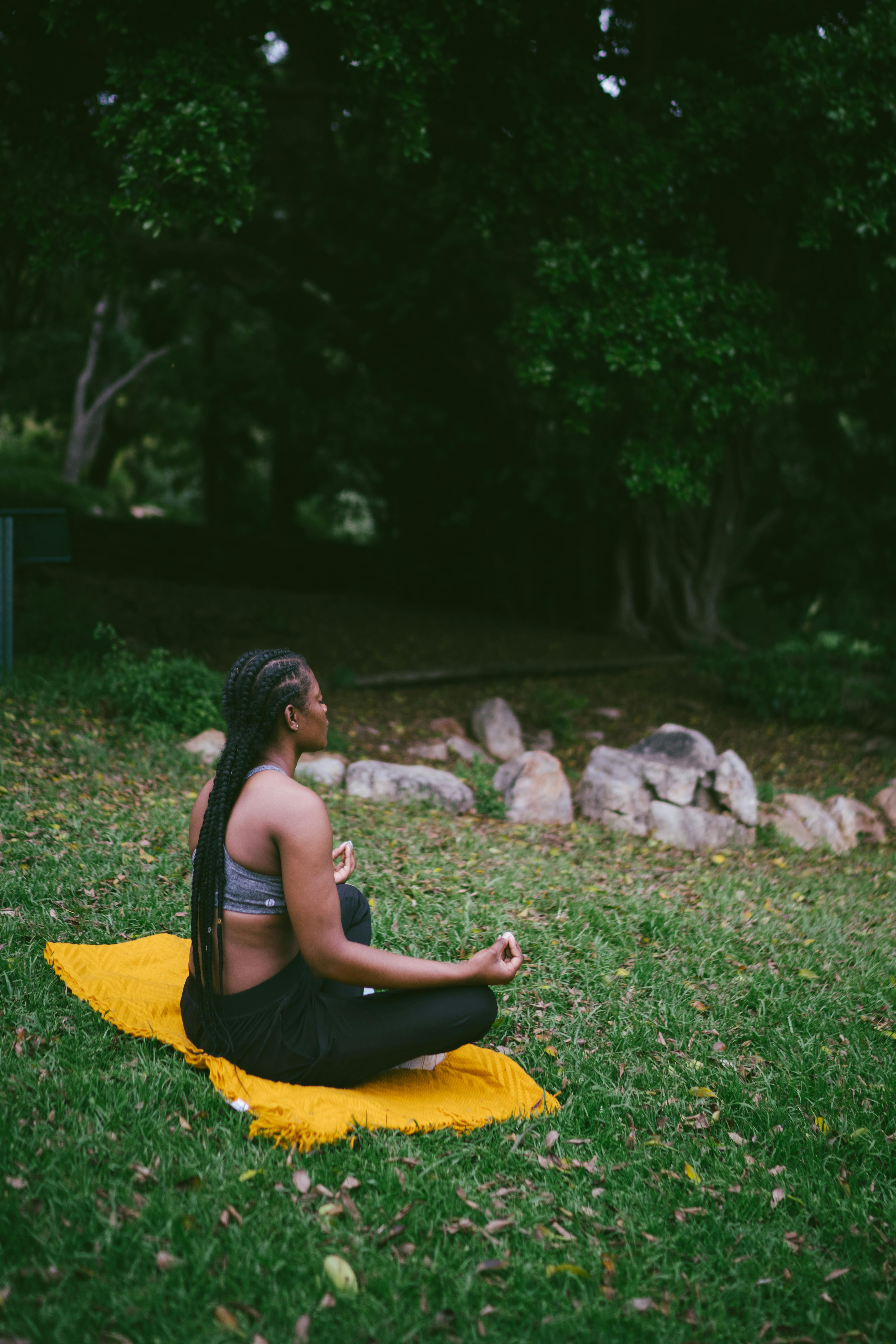 Woman with braided hair meditating cross-legged on a yellow blanket in a grassy park area.