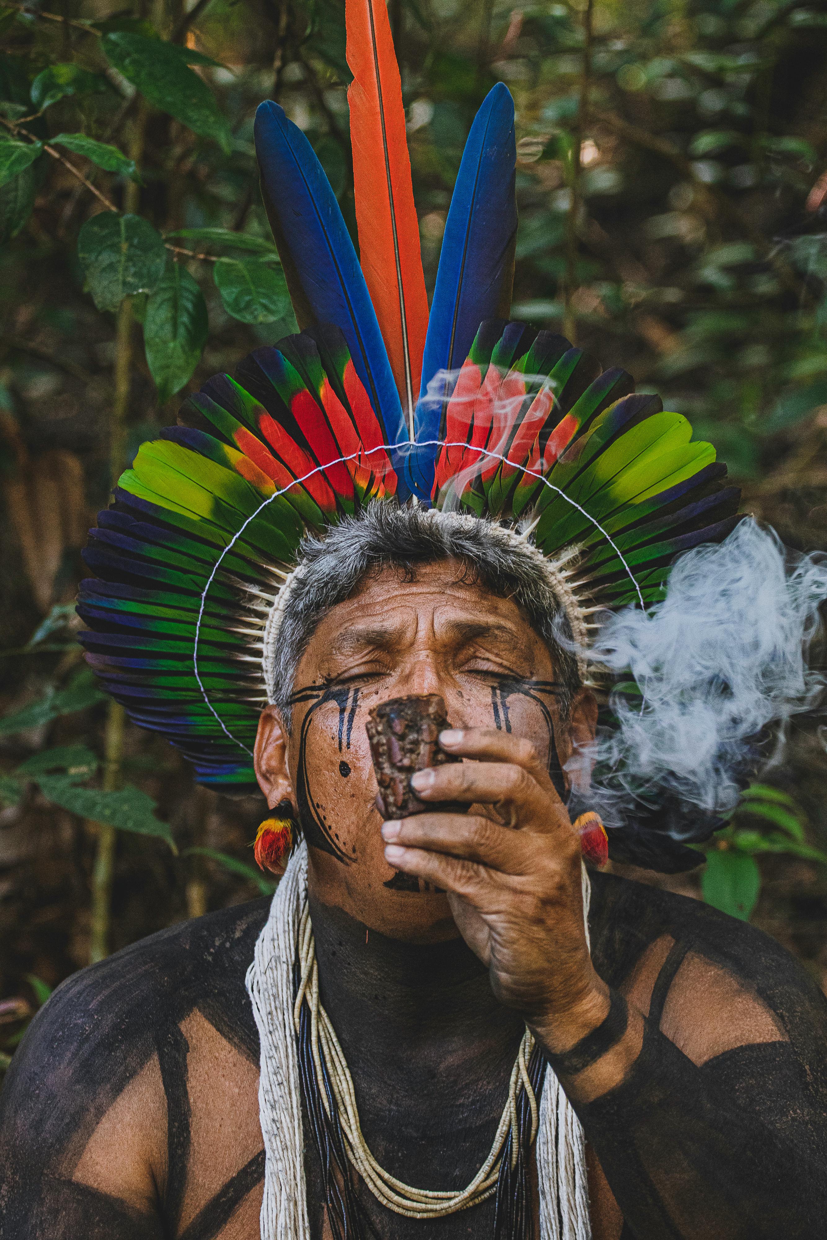 Indigenous man with face paint and a vibrant feathered headdress smoking, surrounded by forest greenery.