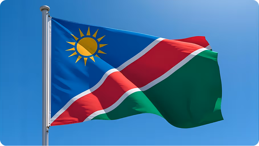 Namibia national flag with a blue field, red diagonal stripe bordered by white, green bottom section, and a golden sun in the upper left corner flying against a clear blue sky.