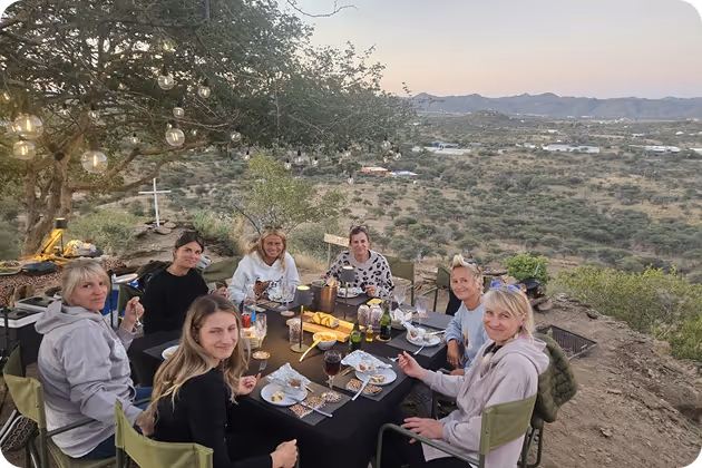 Seven people sitting around a table outdoors on a hill at sunset, enjoying a meal with drinks and string lights hanging above.