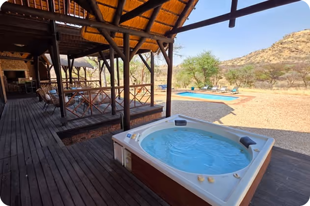 Outdoor wooden deck with a jacuzzi in the foreground, a seating area under a thatched roof, and a swimming pool surrounded by desert landscape with hills and sparse trees.