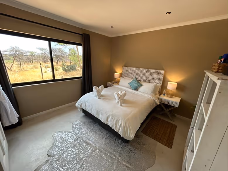 Cozy bedroom with a double bed, white bedding, two bedside tables with lamps, a window showing a dry landscape, and light-colored floor with a decorative rug.