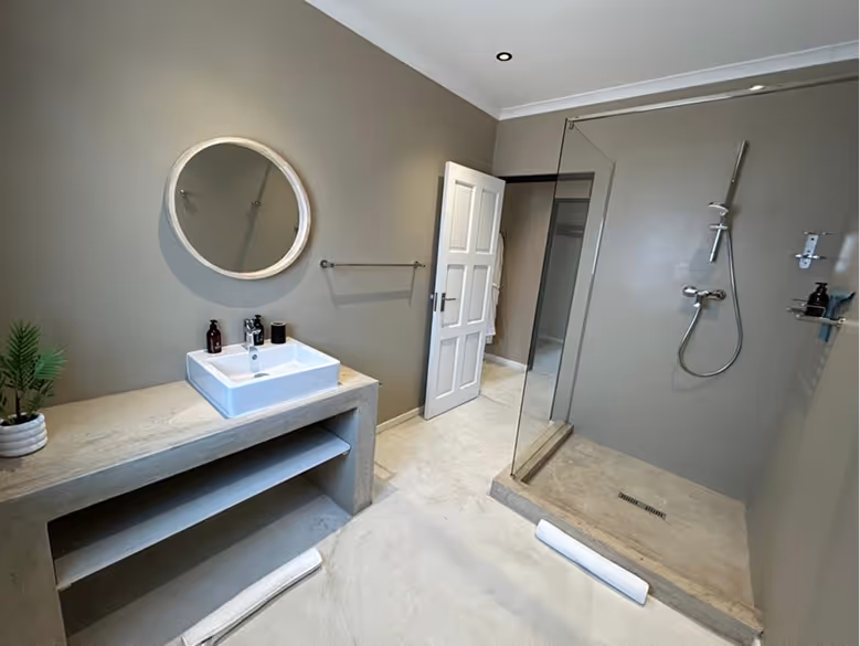 Modern bathroom with beige walls, a white square sink on a stone countertop, round mirror, glass shower enclosure, and an open white door.