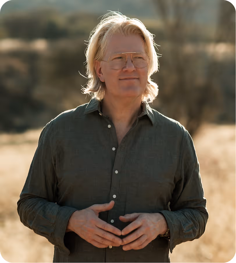 Blond man with glasses wearing a green button-up shirt standing outdoors in a sunlit natural setting.