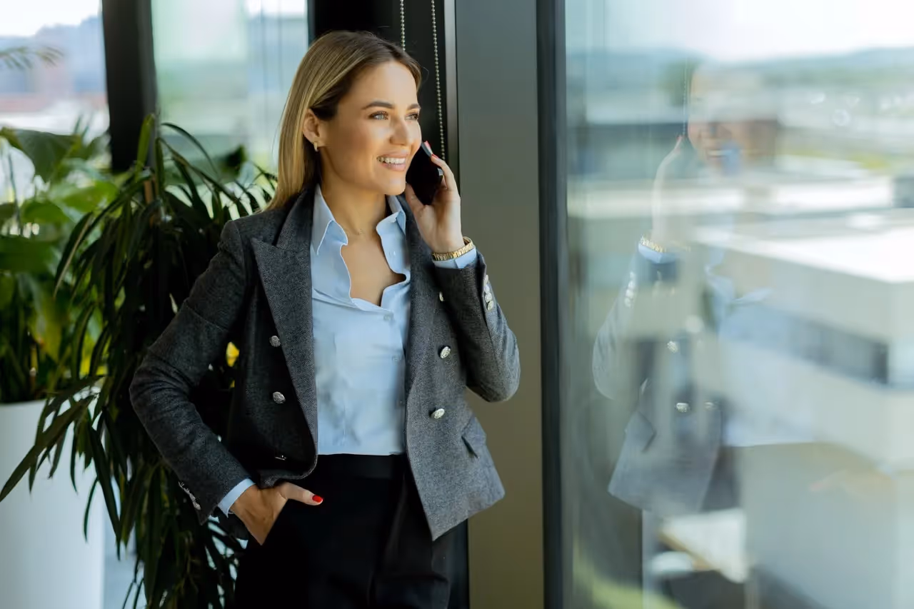 Smiling businesswoman in gray blazer talking on phone while standing by office window with plants in background.