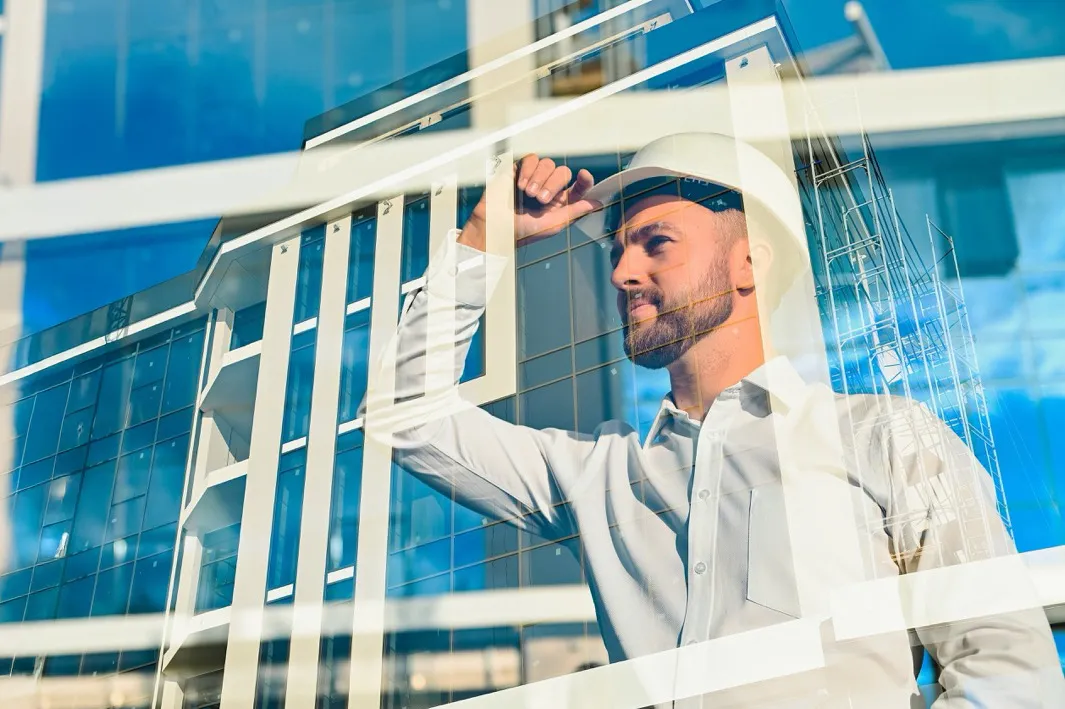 Man in a white hard hat and shirt inspecting a modern glass building under construction with scaffolding.