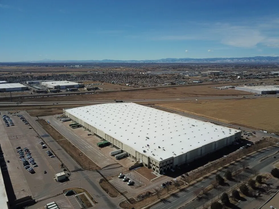 Aerial view of a large white-roofed warehouse surrounded by parking lots and roads with fields and mountains in the background.