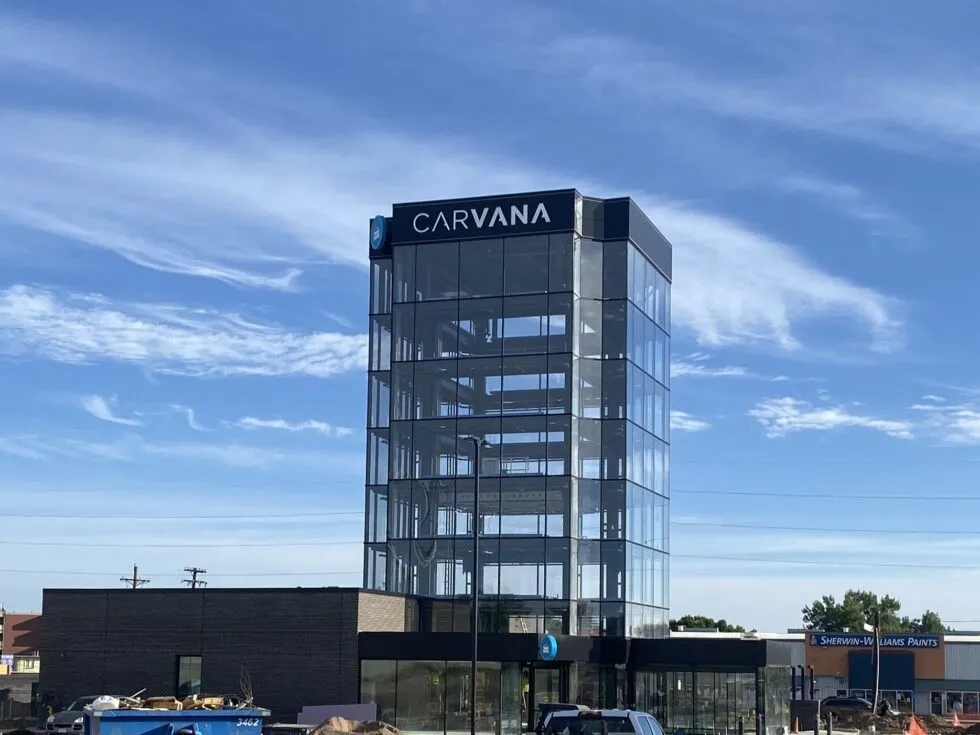 Glass tower with Carvana logo on top against a blue sky, with an adjacent brick building and parked cars.