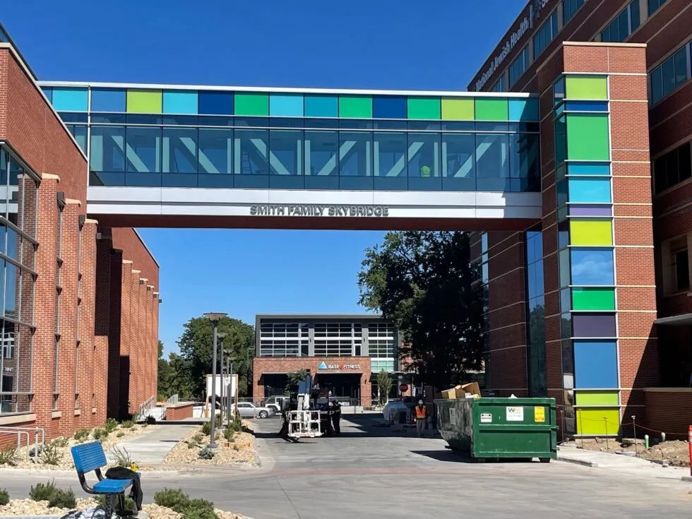 Modern skybridge labeled 'Smith Family Skybridge' connecting two brick buildings with colorful panels, with a clear blue sky background.