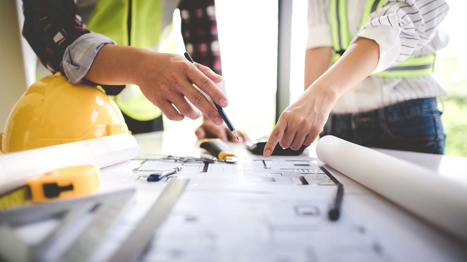 Two people in safety vests reviewing architectural blueprints on a table with a yellow hard hat and measuring tools.