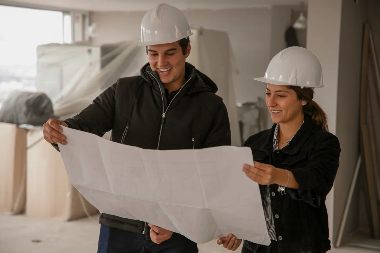 Two architects wearing white hard hats and dark jackets reviewing a large blueprint inside a building under construction.