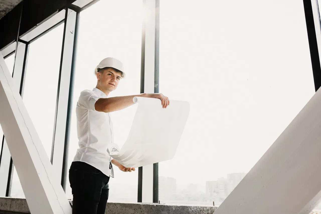 Young male architect or engineer in a white shirt and hard hat holding blueprints in a modern building with large windows.