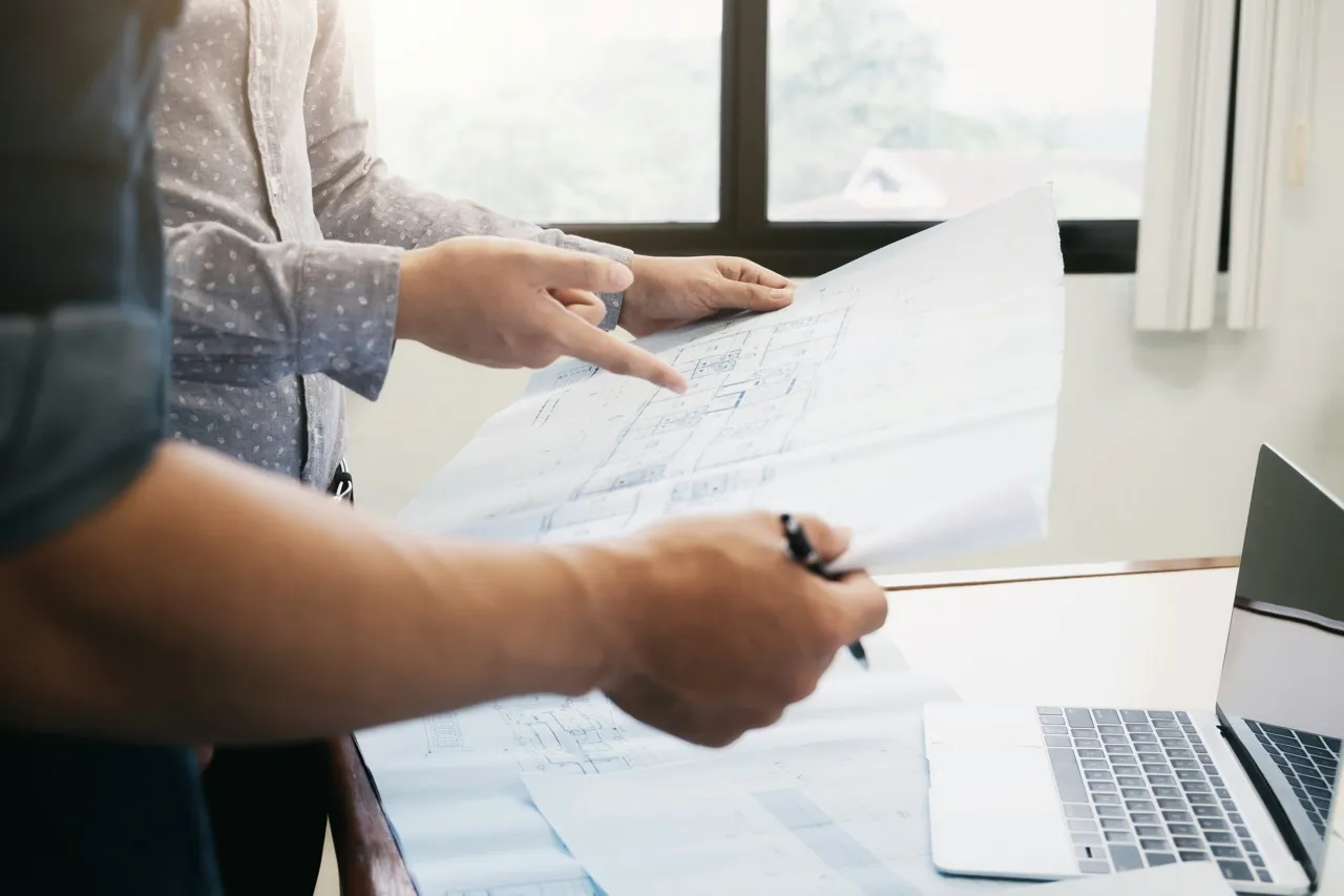 Two people reviewing architectural blueprints together near a laptop on a table.