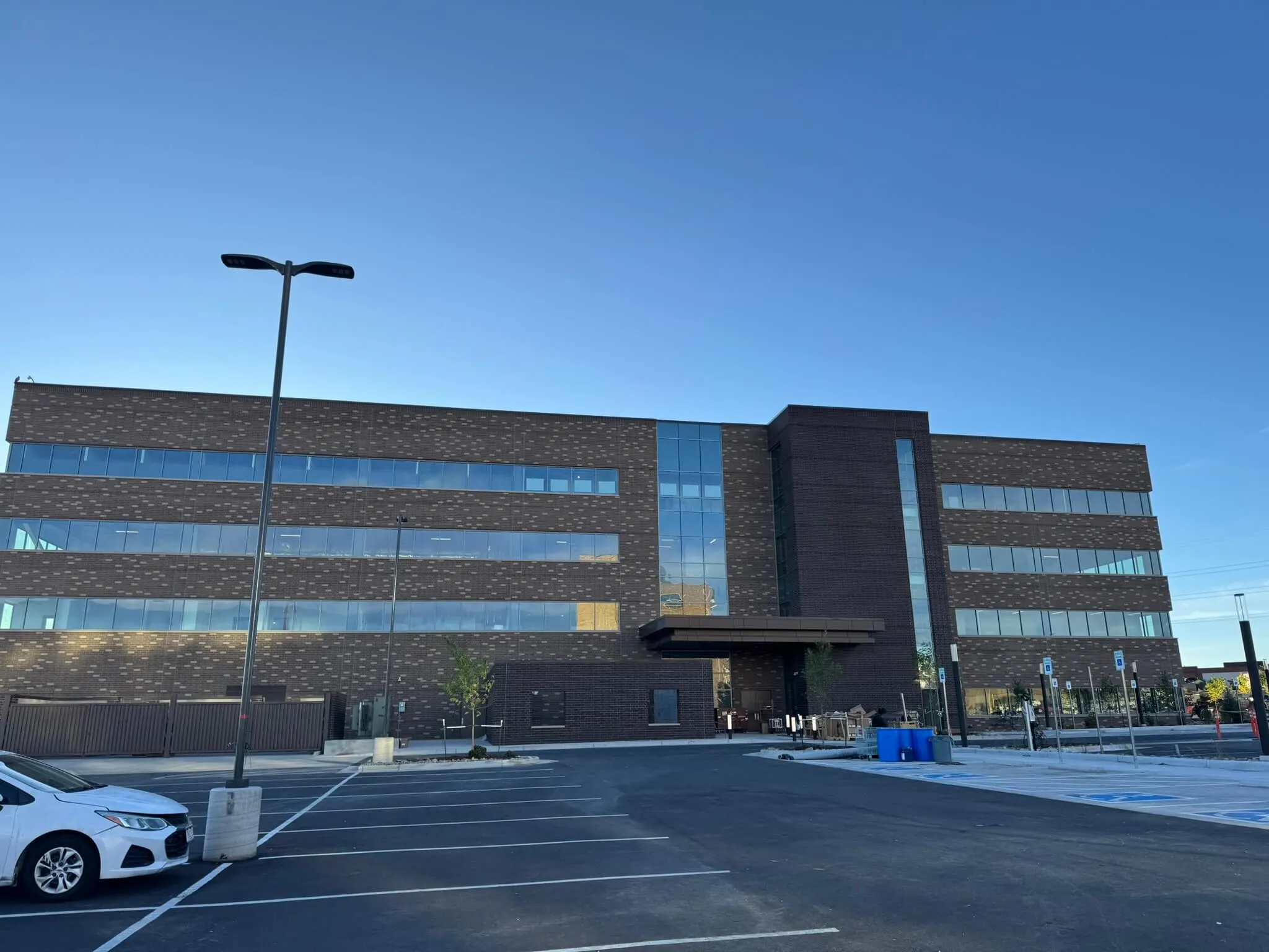 Modern four-story building with dark brick facade and large horizontal windows under clear blue sky, with an empty parking lot in front.