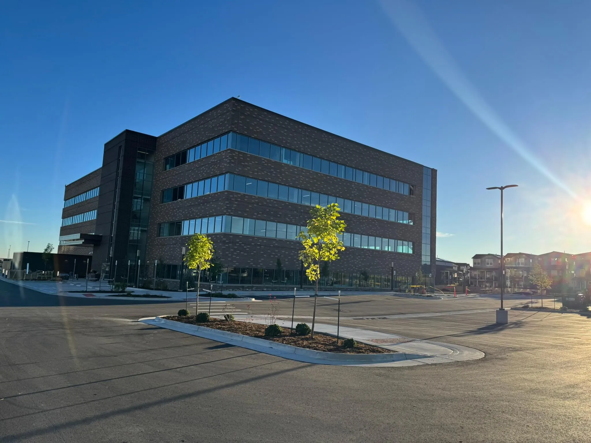 Modern four-story office building with large windows, small landscaped trees, and an empty parking lot under a clear blue sky with sunlight.