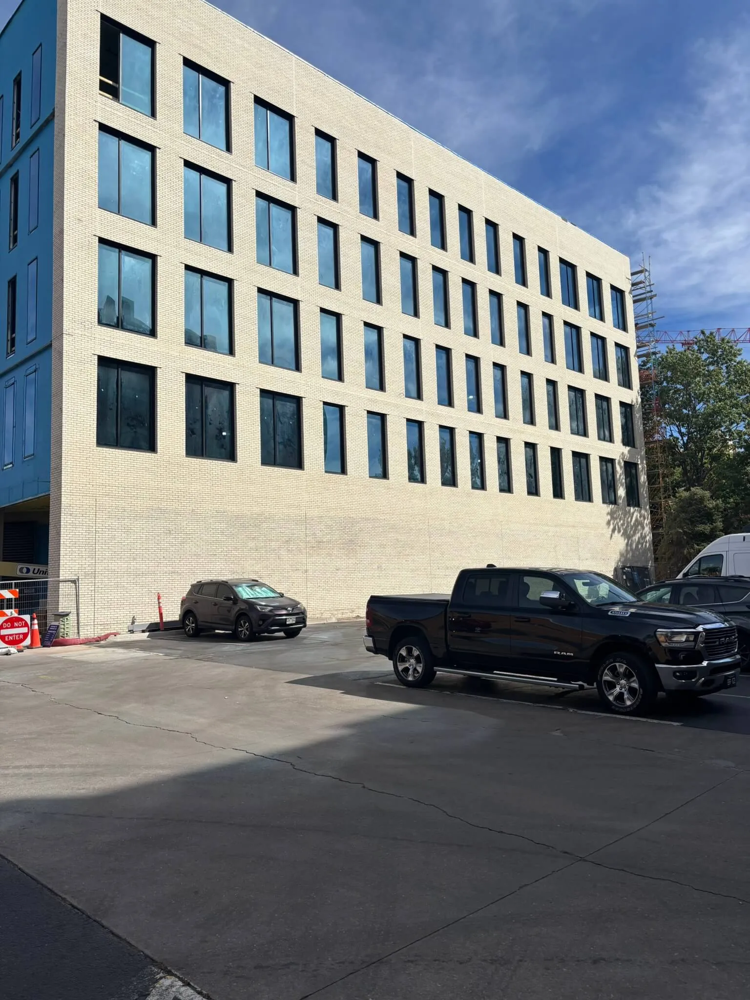 Parking lot with dark pickup truck and SUV beside a beige brick building with multiple rectangular windows under a partly cloudy sky.