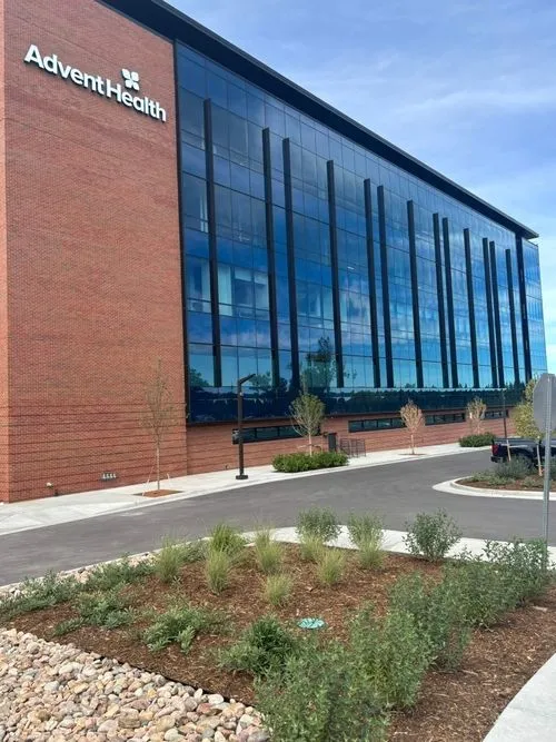 Modern AdventHealth building with reflective glass windows and a brick facade, surrounded by landscaping and a parking area.