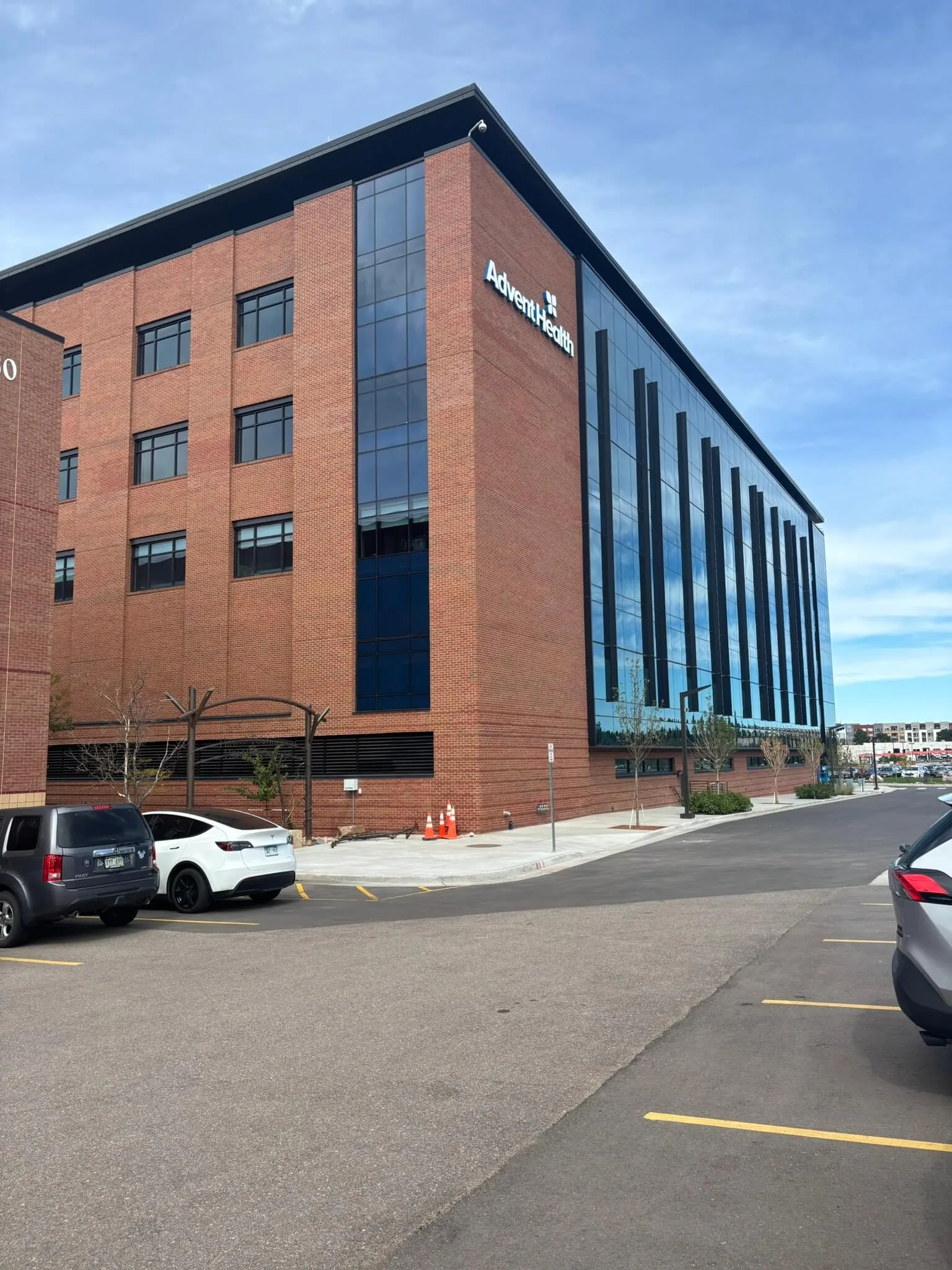 Modern four-story brick building with large glass facade, labeled AdventHealth, under a clear blue sky with parked cars nearby.