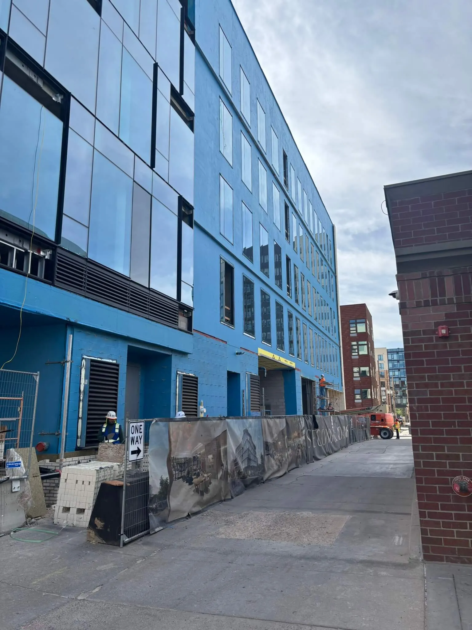 Construction site of a modern blue building with reflective windows, fencing, and workers wearing safety gear.