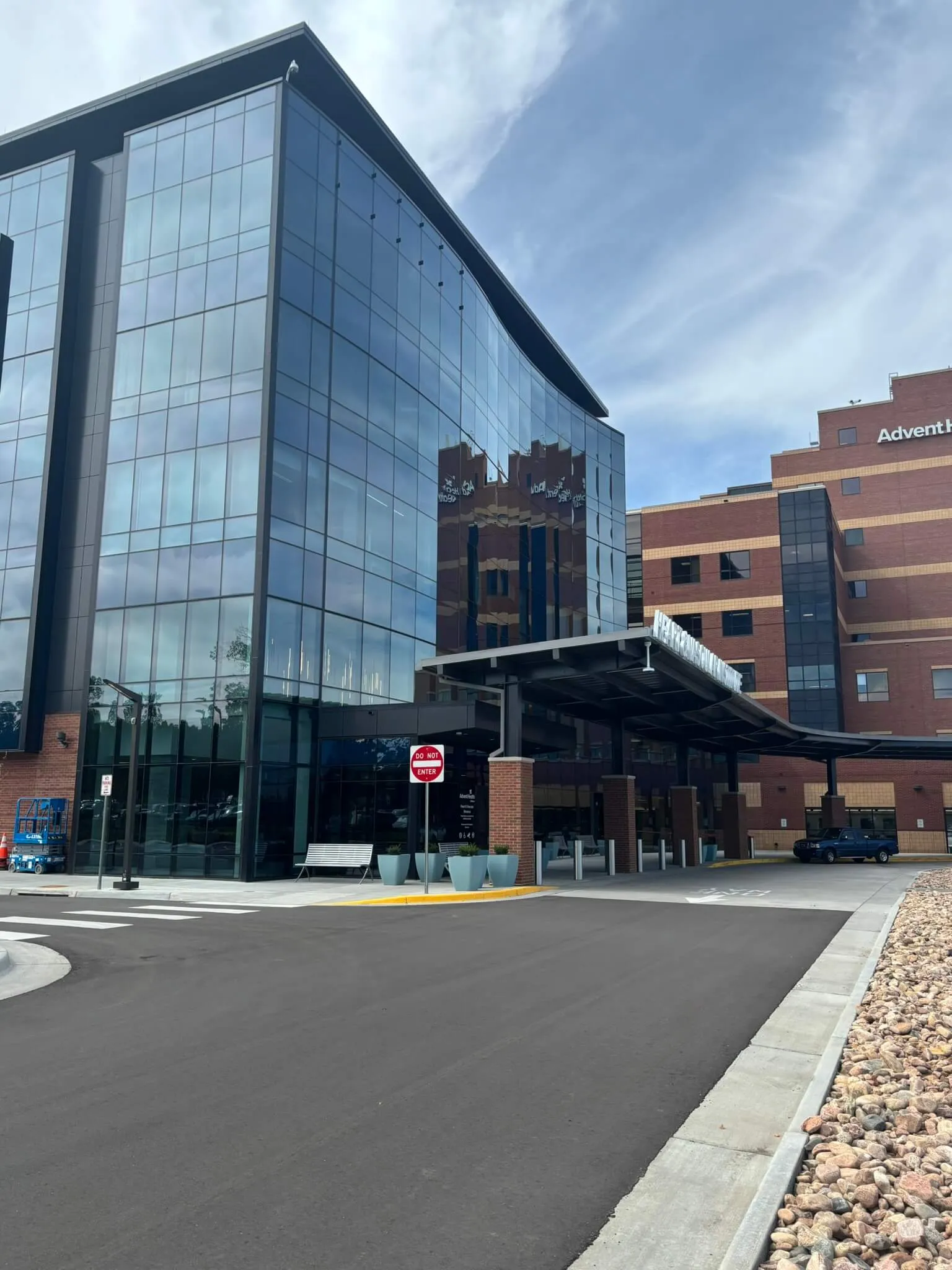 Modern hospital entrance with reflective glass facade, a covered drop-off area, and a no entry sign on a clear day.