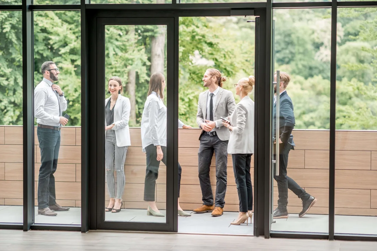 Group of six business professionals casually conversing on an outdoor office entrance with green trees in the background.