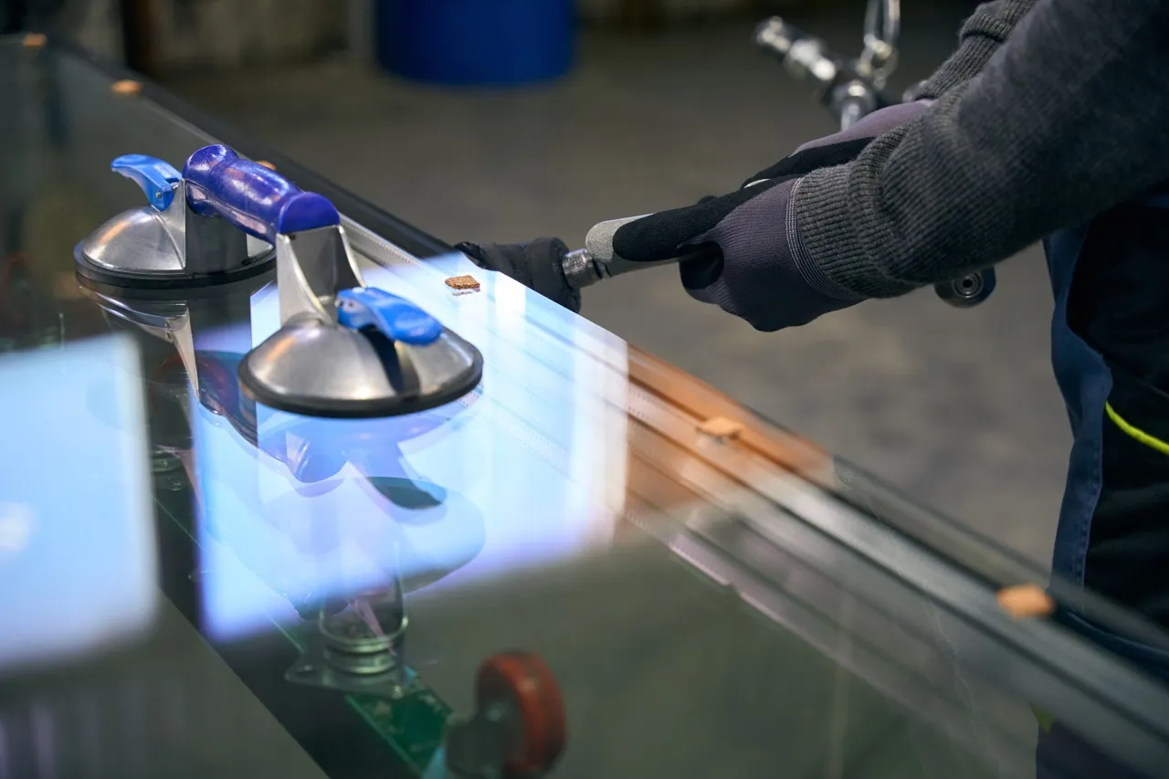 Worker wearing gloves handling a suction cup tool on a sheet of glass in a workshop.