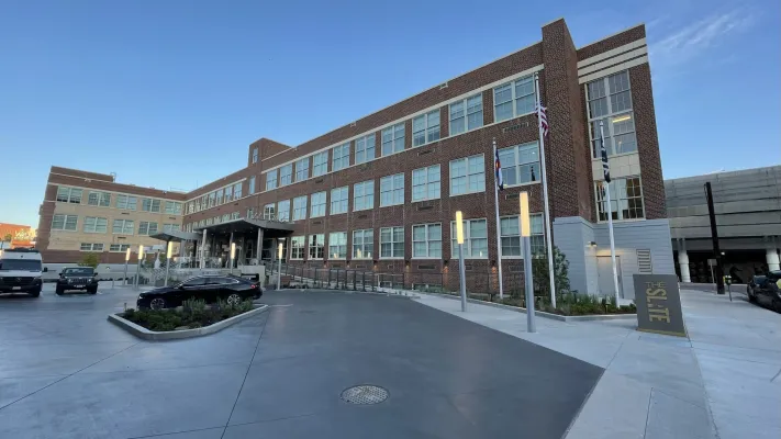 Three-story brick building with large windows and an entrance canopy, surrounded by pavement, cars, and flagpoles under a clear blue sky.