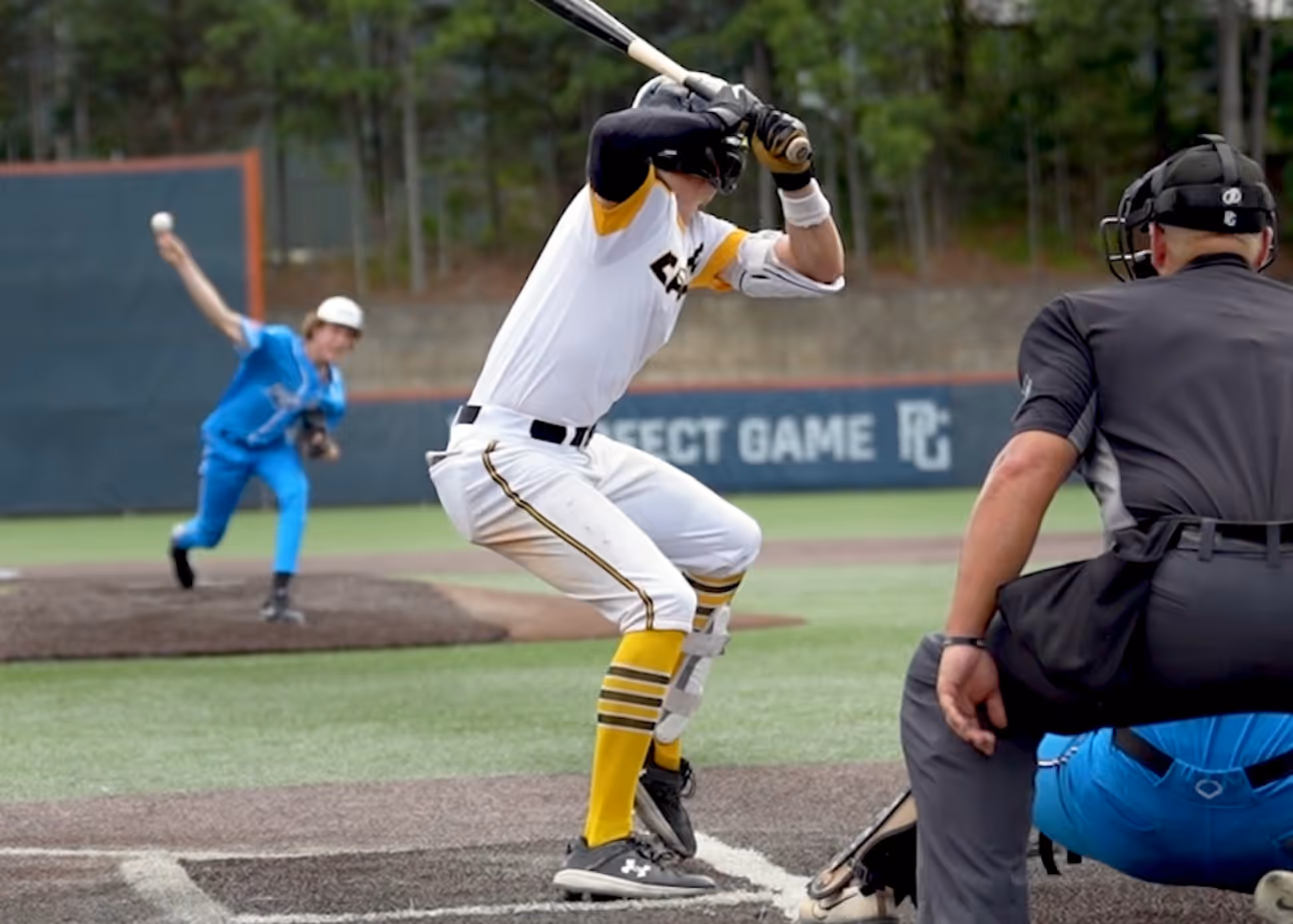 Baseball player ready to swing bat, pitcher in the background throwing ball.