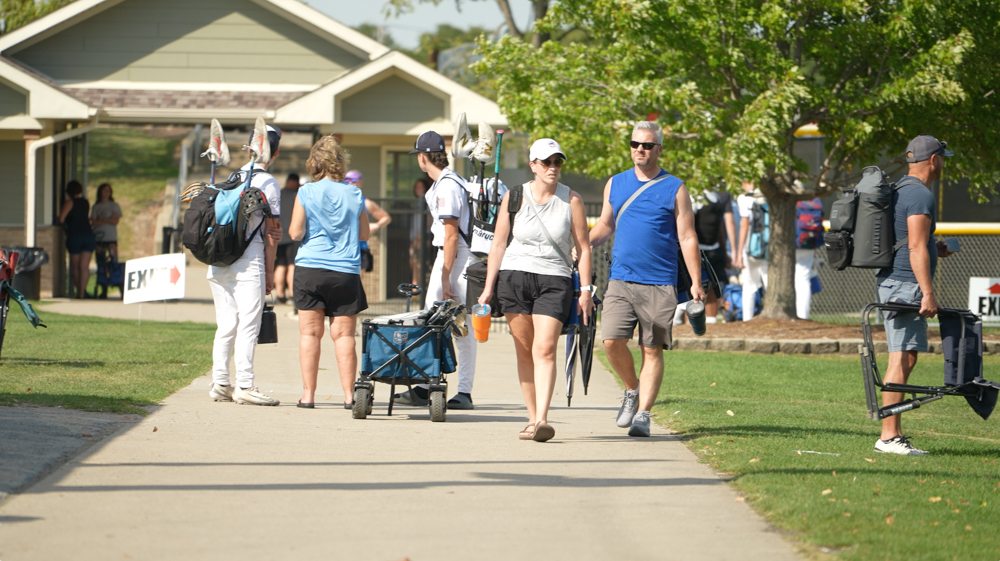 People walking on a paved path on a sunny day near a baseball area, some carrying baseball bags and equipment.