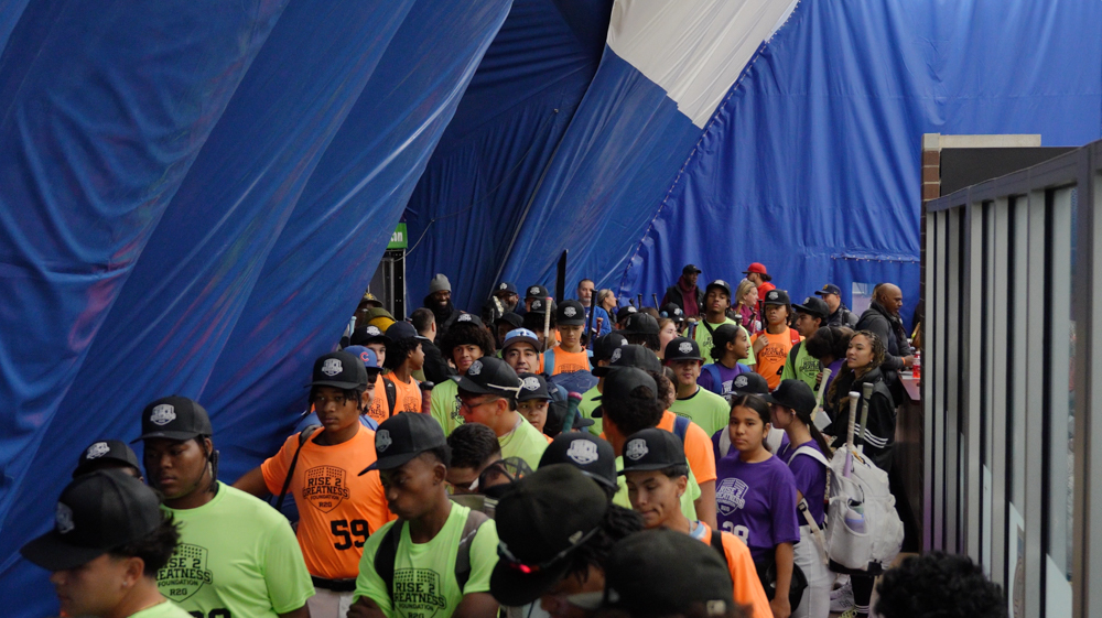 Group of young baseball players wearing colorful jerseys and black caps standing under blue tent fabric at an event.