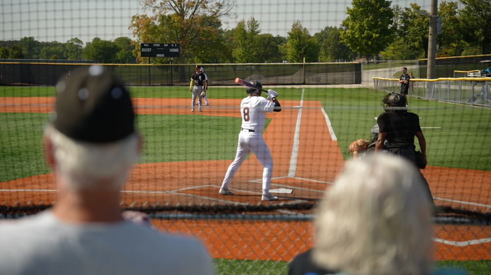 Baseball batter standing ready to hit while catcher and umpire crouch behind home plate, viewed from behind two spectators through protective netting.
