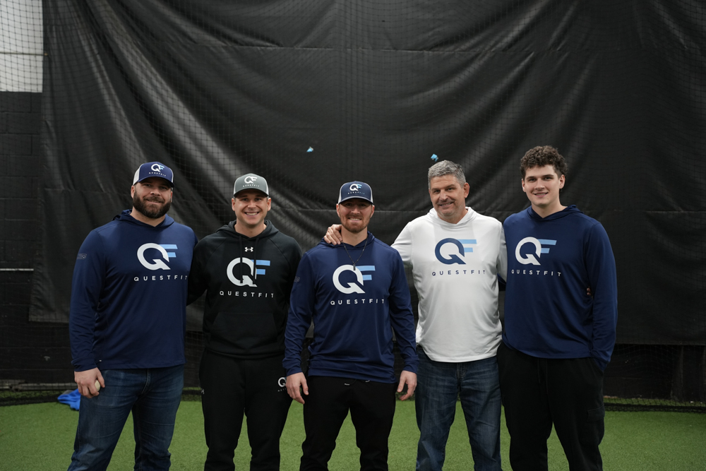 Two men wearing matching QuestFit caps and shirts sit at a black QuestFit table with laptops at an outdoor baseball event.