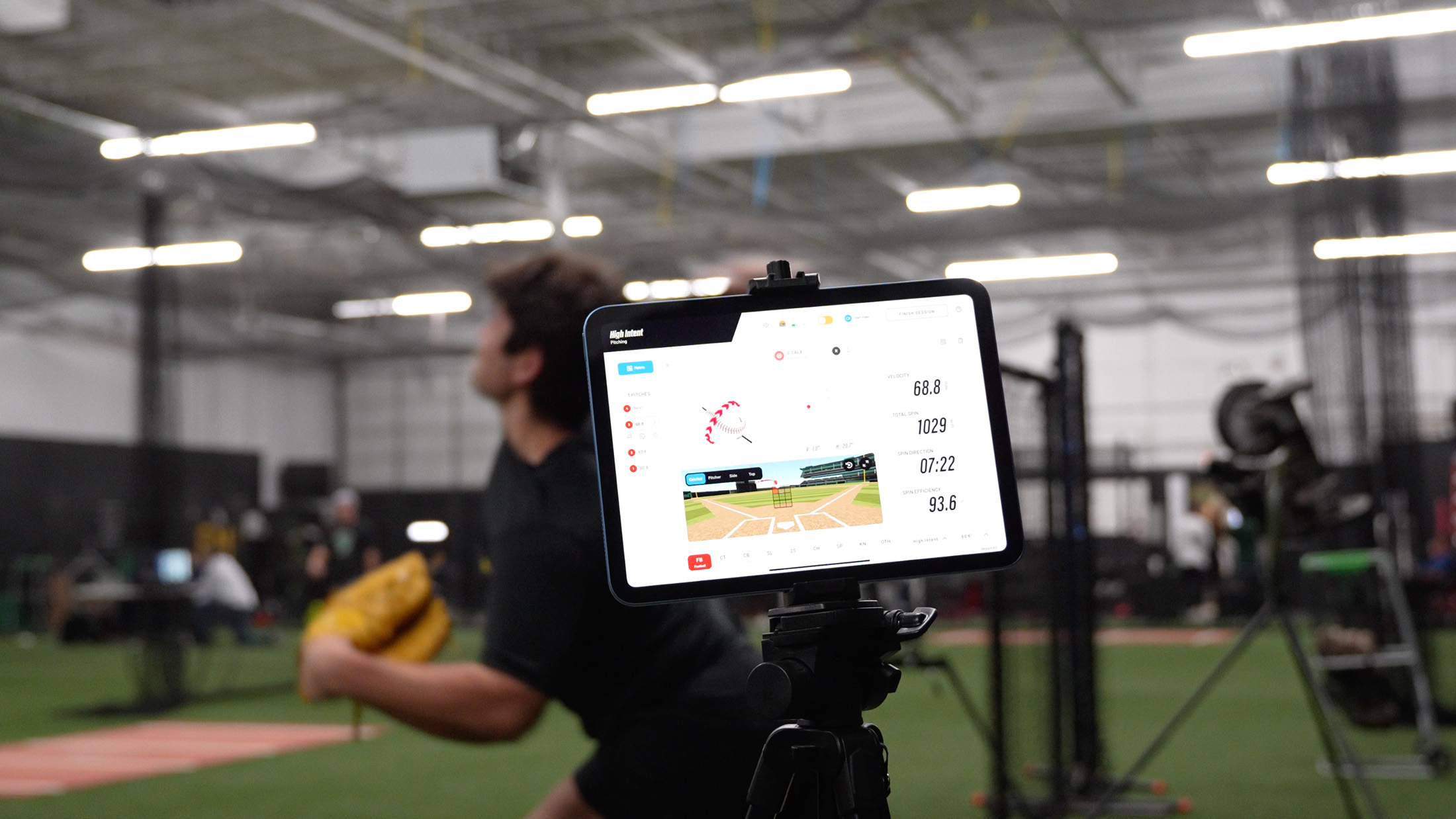 People walking on a paved path on a sunny day near a baseball area, some carrying baseball bags and equipment.