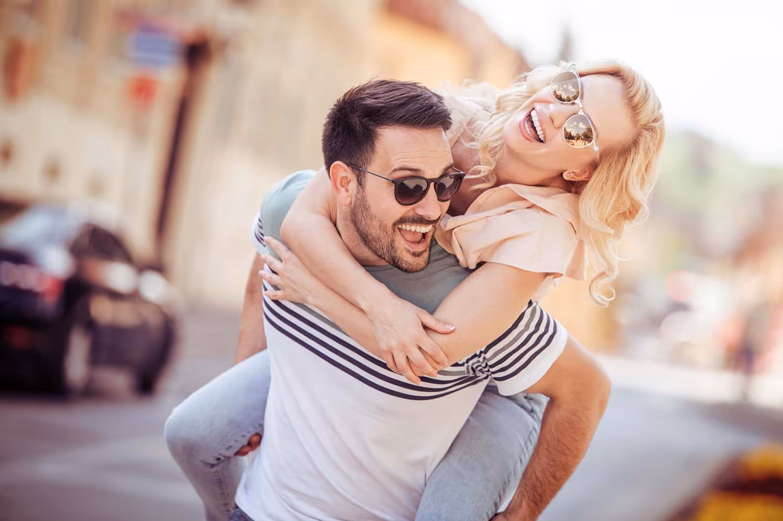 Smiling man wearing sunglasses giving a piggyback ride to a laughing woman also in sunglasses on a sunny street.