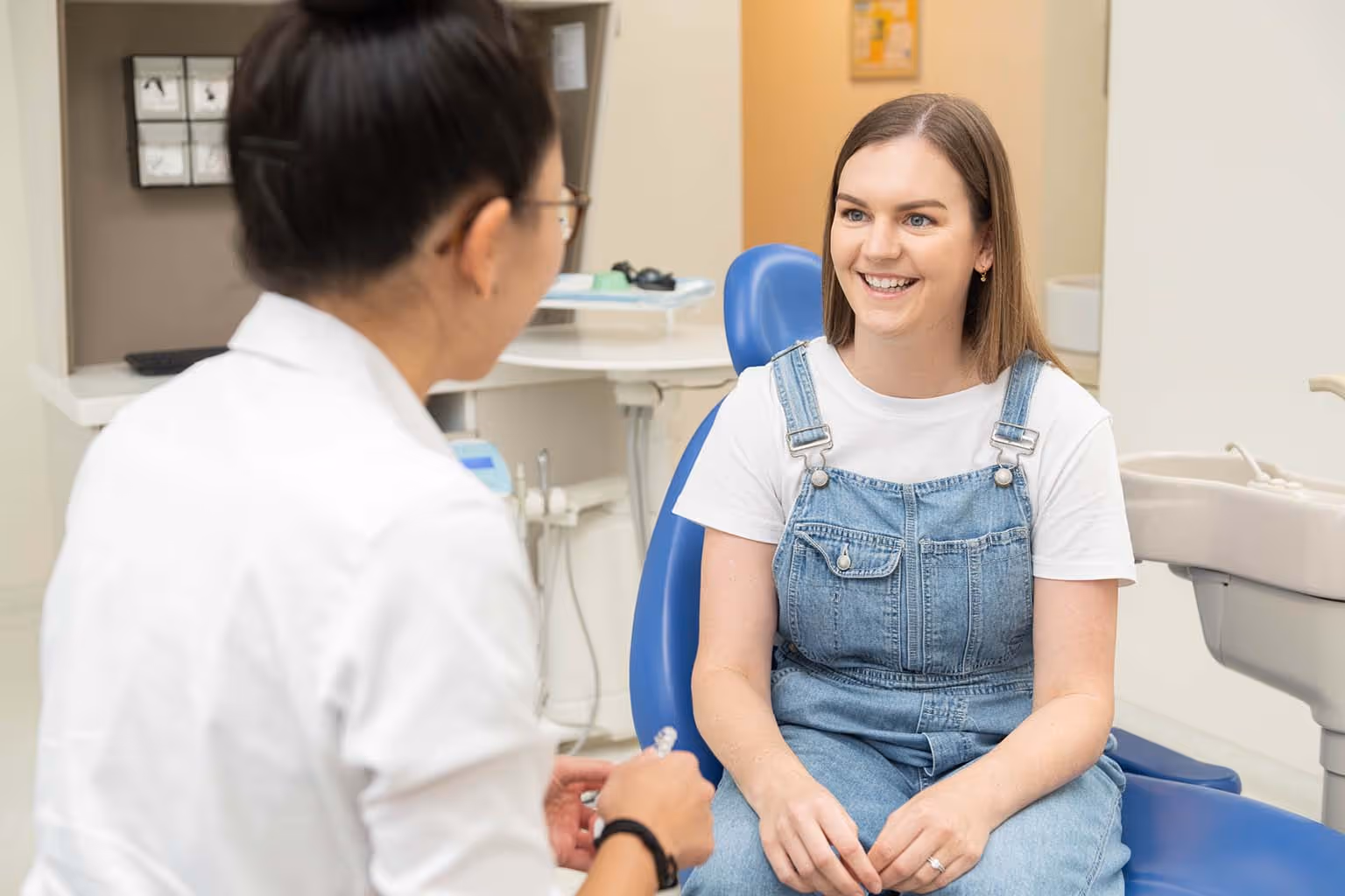 Smiling woman in denim overalls sitting in a dental chair talking to a female dentist.