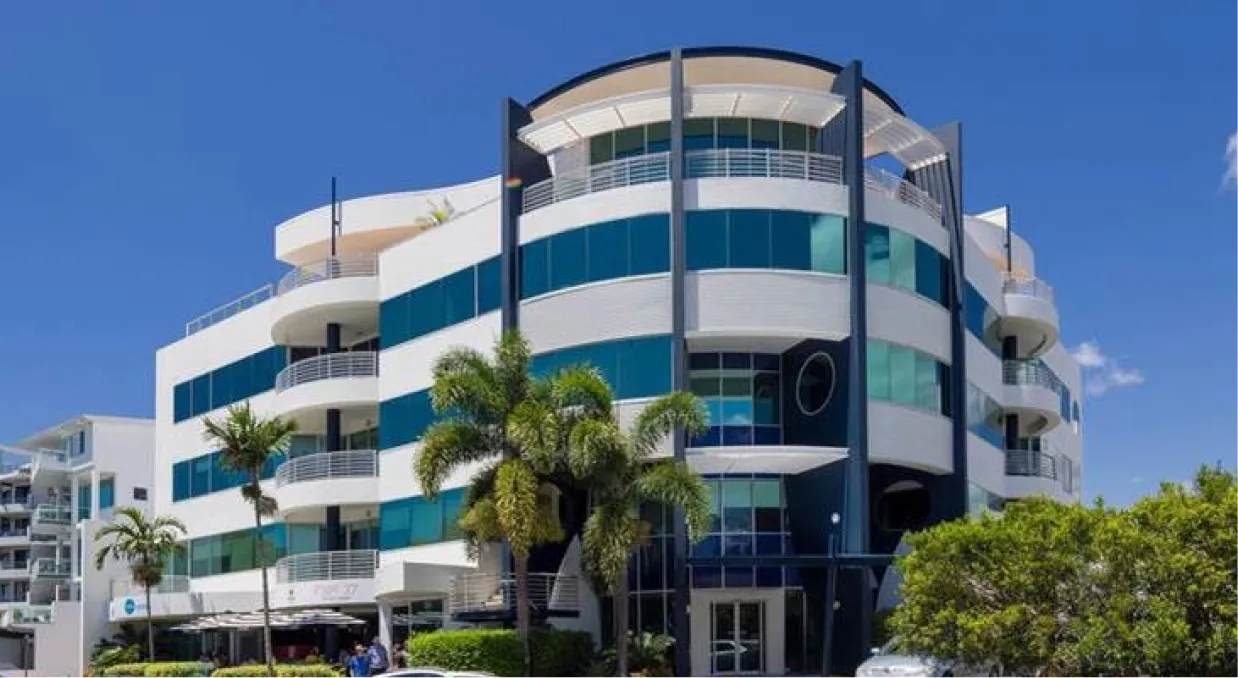 A large white building with balconies and blue tinted windows surrounded by green trees and blue sky.