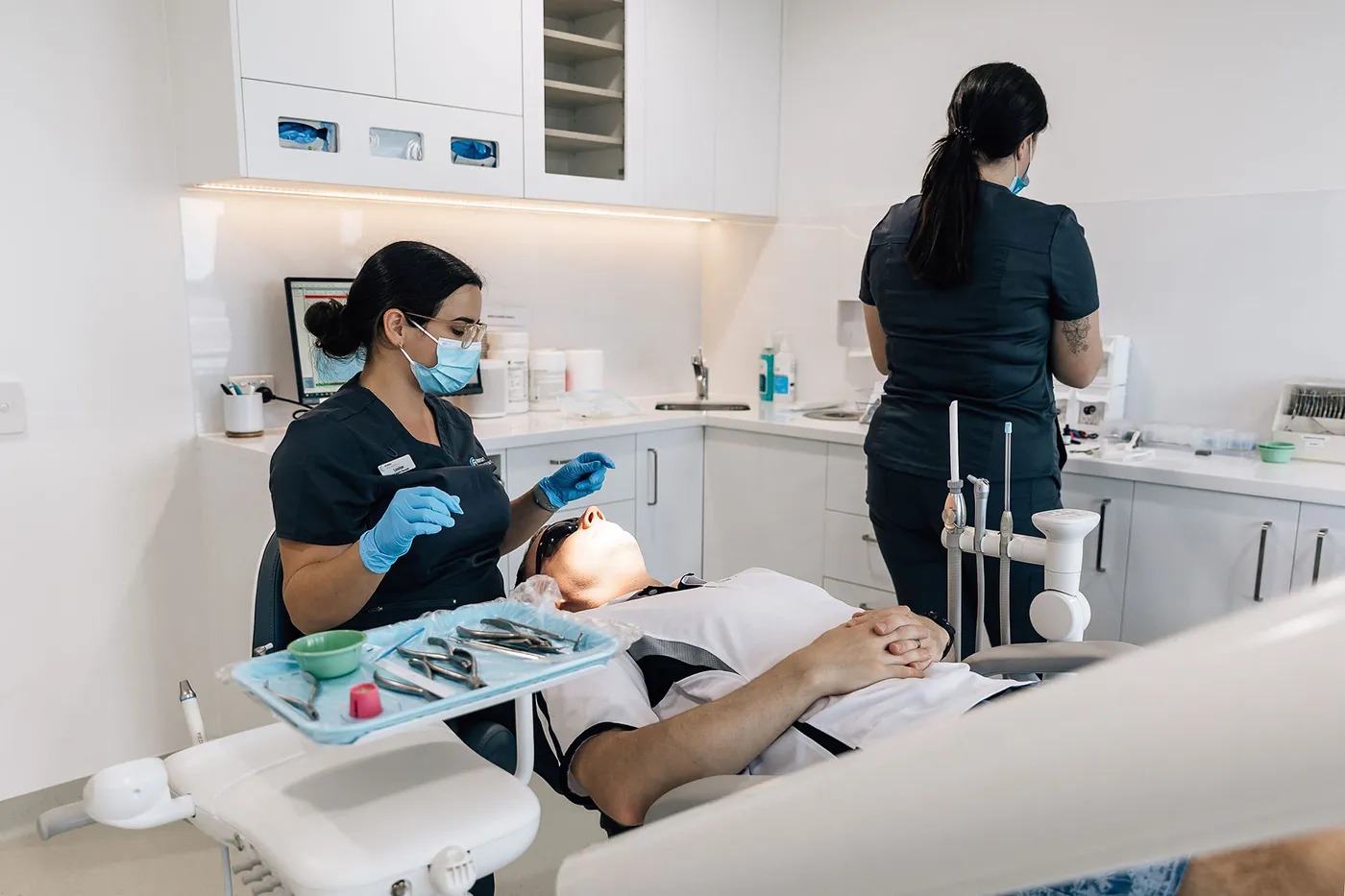Dentist wearing a mask and gloves preparing to treat a patient lying back in a dental chair in a modern clinic.