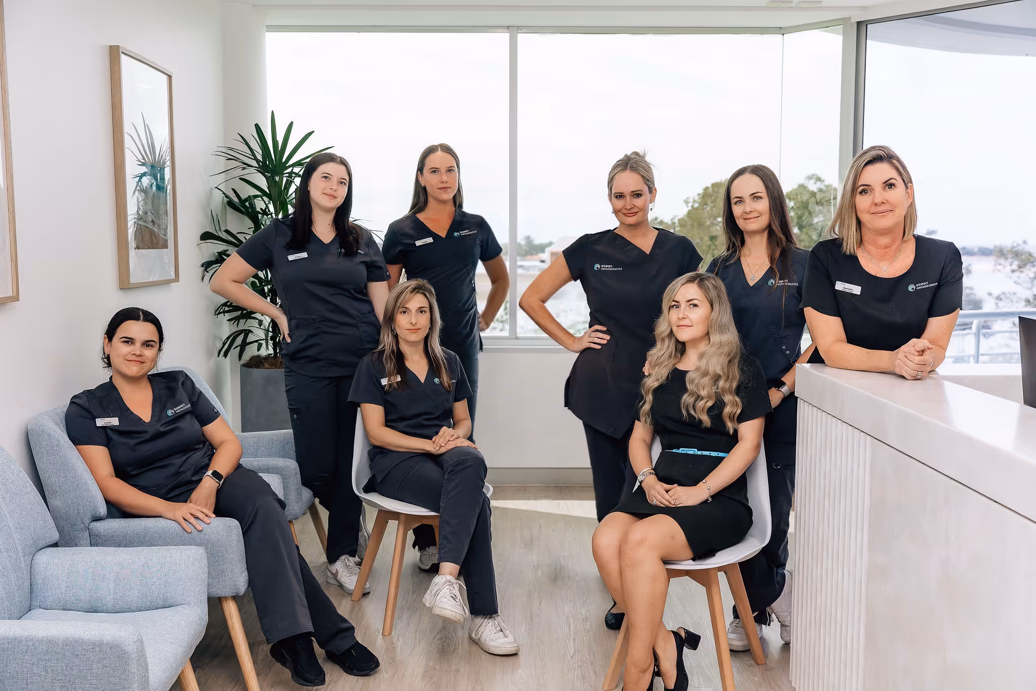 Seven women in dark medical scrubs posing in a modern, bright office with large windows and plants.
