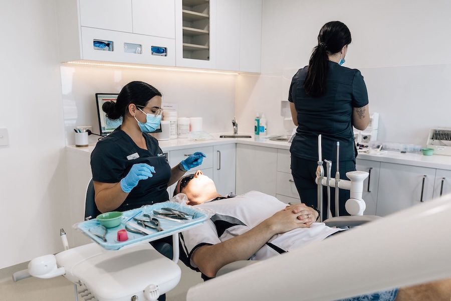 Dentist wearing a mask and gloves preparing to examine a patient reclining in a dental chair while a dental assistant stands at the counter.