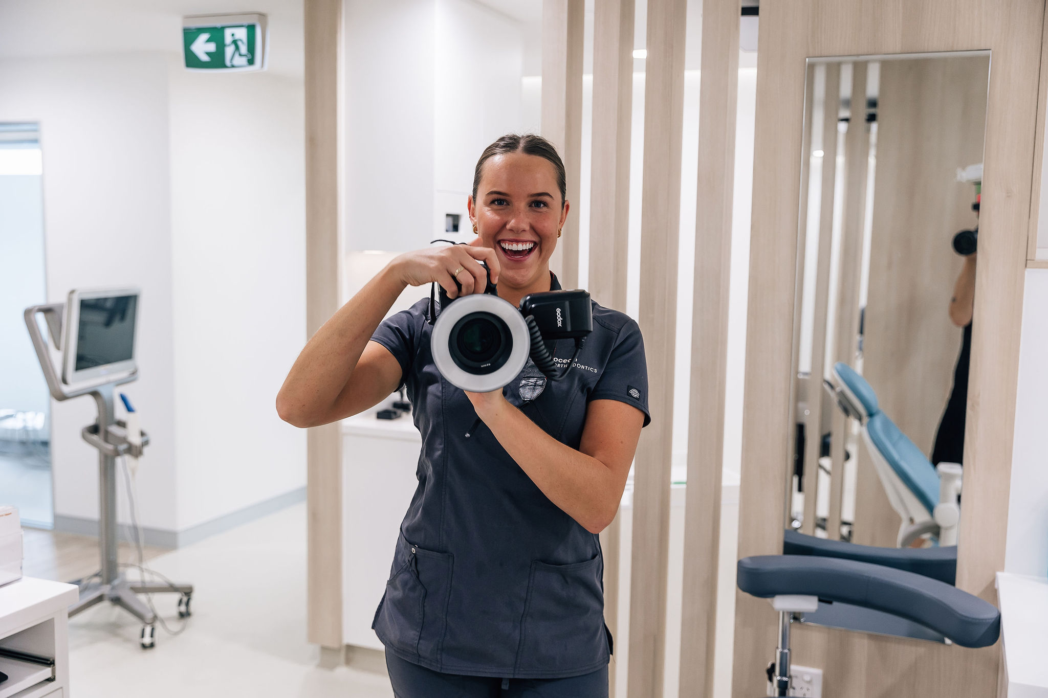 A woman in a dentist surgery, holding a camera and smiling.