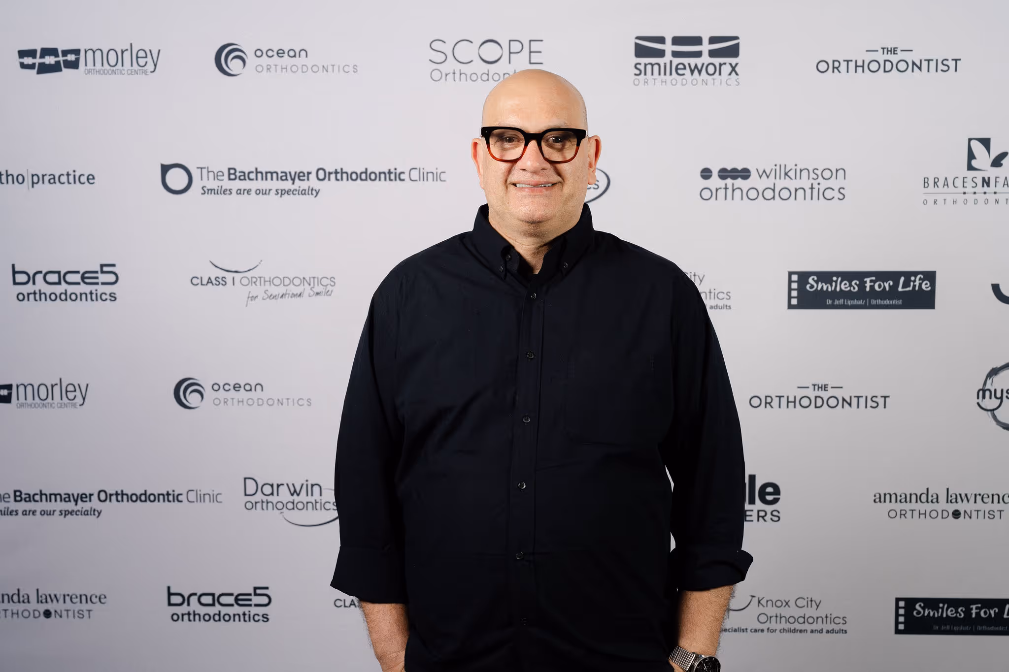 Smiling bald man wearing glasses and a black shirt standing in front of a white backdrop with orthodontics clinic logos.