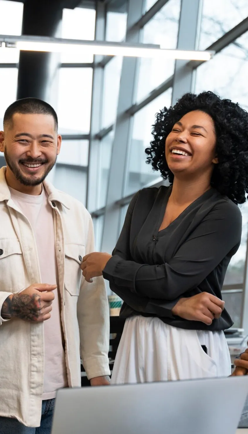 Two colleagues smiling and laughing together in a modern office with large windows.