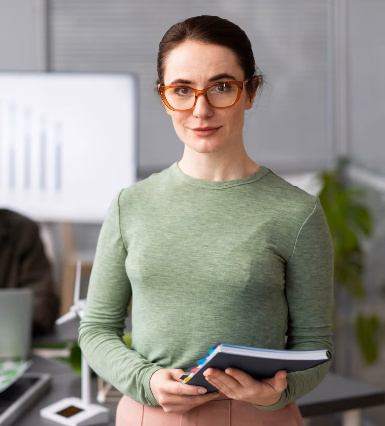 Woman with brown hair and orange glasses wearing a green long-sleeve shirt holding a notebook in an office setting.