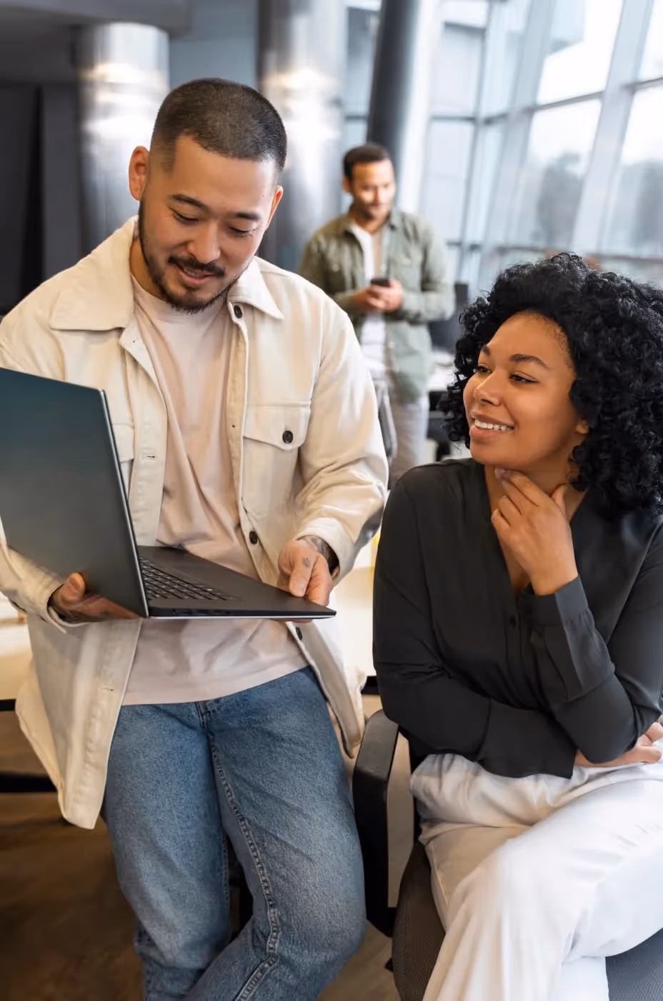 Smiling man showing content on laptop to a seated woman in a modern office with a man using a smartphone in the background.